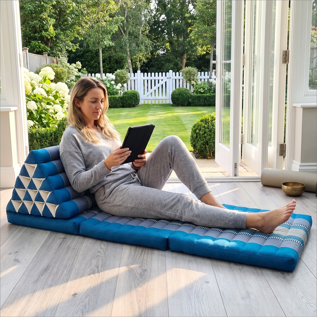 Woman reclining with a tablet on a blue Jumbo Thai triangle floor mattress opened out as a lounger, in a garden sunroom
