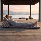 Woman lying back, hands behind her head, on a Mix Elephant Jumbo Thai triangle floor mattress opened out flat, on a lakeside pavilion at dawn