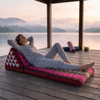 Woman lying back, hands behind her head, on a pink Jumbo Thai triangle floor mattress opened out as a lounger, on a lakeside pavilion at dawn