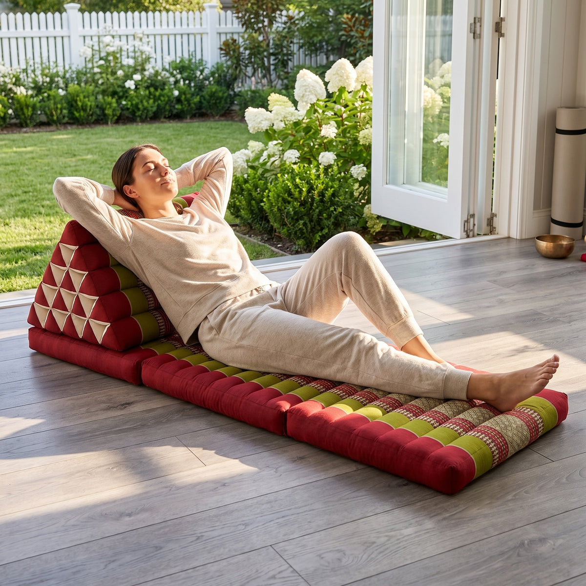 Woman lying back, hands behind her head, on a red and green Jumbo Thai triangle floor mattress opened out as a lounger, in a garden sunroom